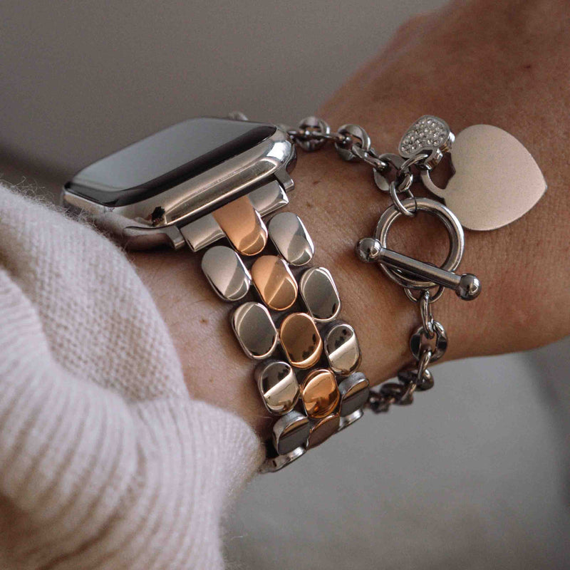  A gold stainless steel Apple Watch band styled on a wrist, paired with other silver and charm bracelets.